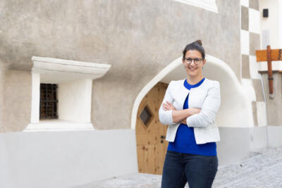 Caty Emonet steht mit leuchtend blaum Obwerteil und weisser Weste darüberlächelnd vor einem alten Engadiner Haus